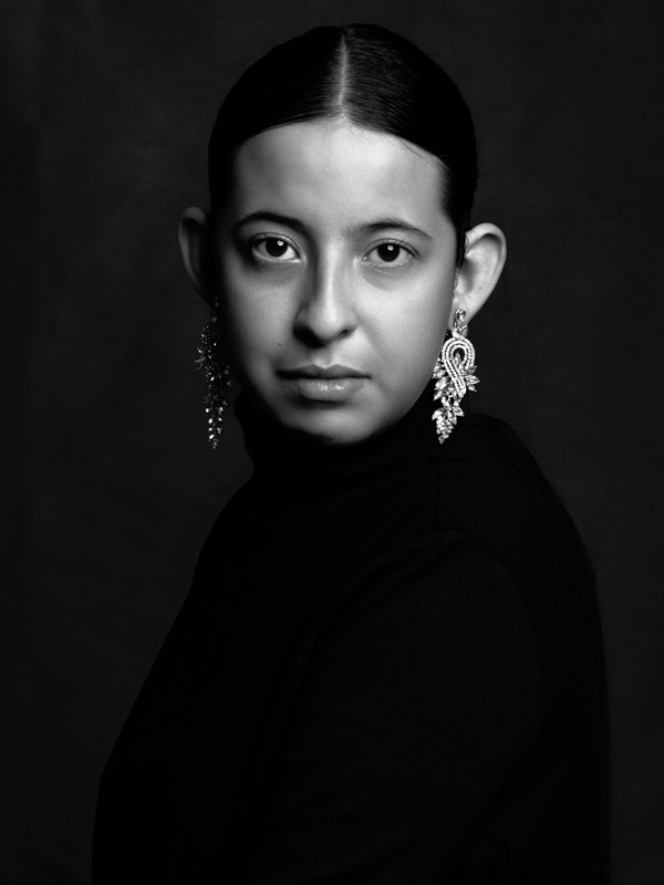 black & white; a tight portrait of a beautiful young woman wearing a black turtleneck, diamond earrings. Background is dark. 