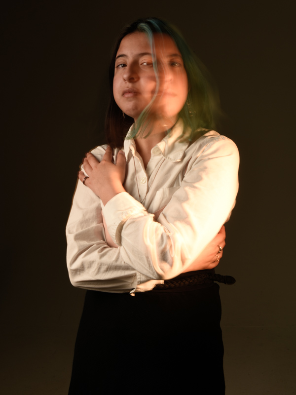 color; Beautiful young girl wearing a white long sleeve shirt and black skirt. She is projecting a second face with light painting technique. Background is dark and moody. 