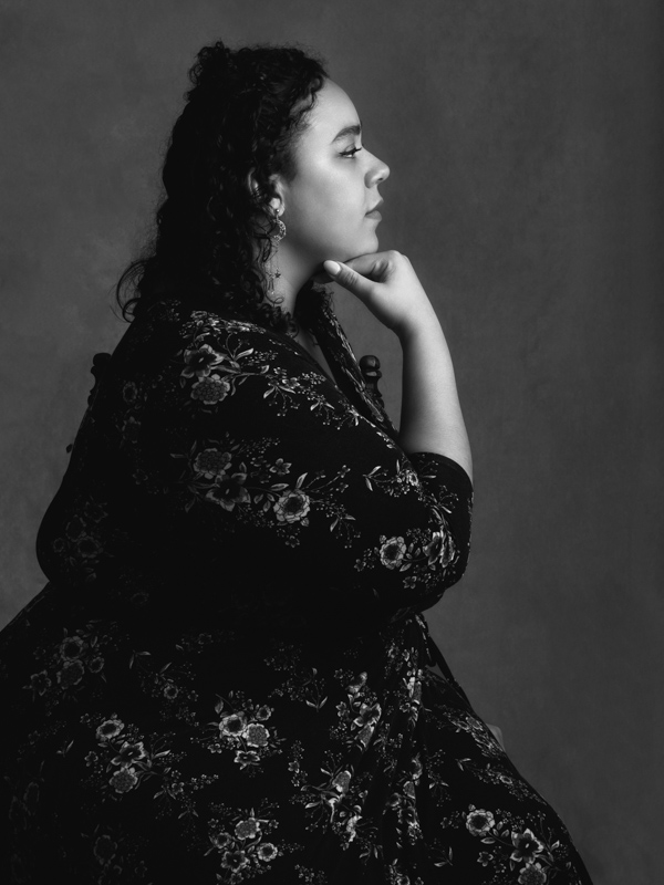 black & white: A profile portrait of a beautiful woman, sitting, with her right hand under her chin. Dress is black with flower pattern. Background is a lighter tone. 