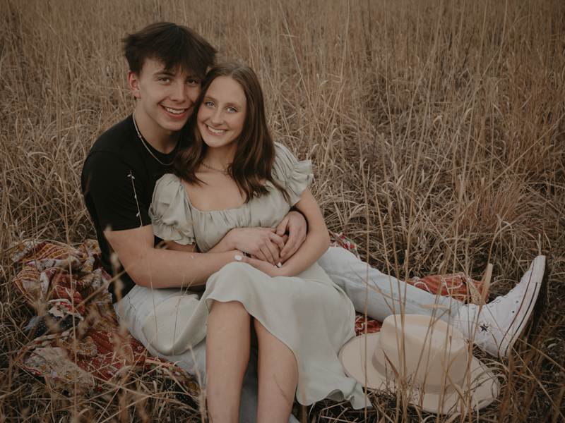 Couple shot of the two sitting on a blanket in a field both looking at camera