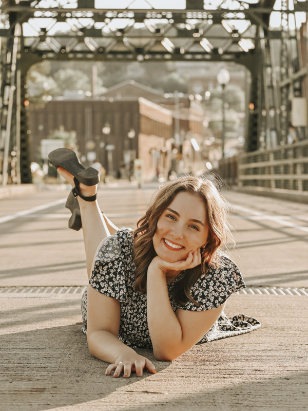 Highschool senior portrait of girl smiling lying on her stomach on a road under a green bridge